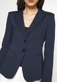 Granatowa, dopasowana marynarka wykonana z gładkiej tkaniny, z klapą z wycięciem, dwoma przednimi kieszeniami i dwoma czarnymi guzikami.