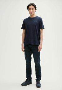 Tricou din bumbac de culoare bleumarin, cu mâneci scurte și gât rotund. Prezintă un logo brodat pe piept. Este combinat cu blugi din denim închis și adidași negri.