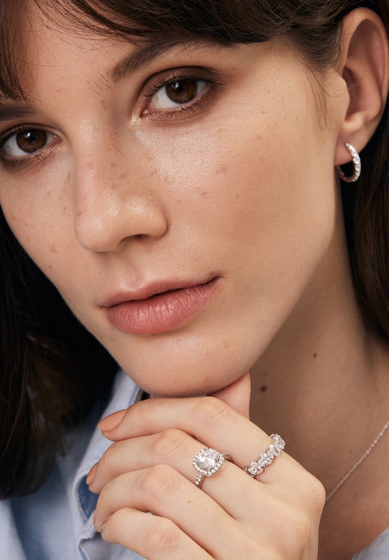 Trzy srebrne pierścionki z przezroczystymi kamieniami, z okrągłym kamieniem centralnym otoczonym mniejszymi, oraz teksturowany srebrny kolczyk w kształcie obręczy.