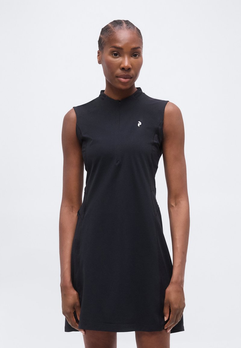 Γυναίκα με κοτσίδες που φοράει ένα μαύρο αμάνικο φόρεμα με μικρό λευκό λογότυπο στο στήθος, στέκεται μπροστά σε ένα απλό ανοιχτό φόντο.