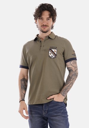 Uomo con capelli ricci e tatuaggi su entrambe le braccia indossa una polo verde oliva con patch in stile militare e jeans blu, in piedi con una mano in tasca.