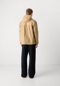 Jasnobeżowa kurtka z kapturem, z elastycznymi mankietami, luźny krój i prosty design, w połączeniu z ciemnymi spodniami typu wide-leg oraz brązowym obuwiem.