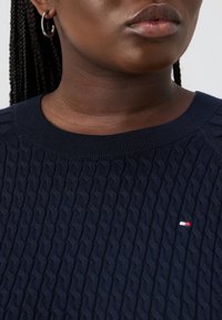 Laivastonsininen neulepaita, jossa on teksturoitu palmikkokuvio, ribbireunainen pyöreä pääntie ja pieni logo punaisia, valkoisia jasinisiä värejä.