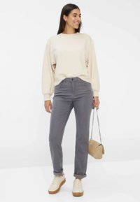 Beige Sweatshirt mit überschnittenen Schultern, graue Slim-Fit-Hose und cremefarbene Sneaker. Das Modell hält eine kleine beige Tasche mit einer Kettenriemen.