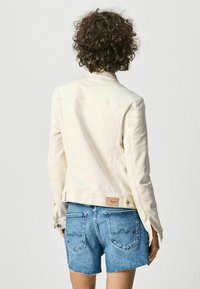Light beige denim jacket with long sleeves, fitted design, and button closures at the cuffs, worn over blue denim shorts with frayed hems.