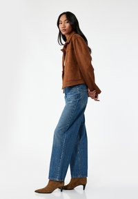 Ruskea nubukka takki, jossa on kaulus ja kaksi etutaskua, yhdistettynä vaaleansinisiin leveälahkeisiin farkkuihin ja vaaleanruskeisiin teräväkärkisiin nilkkasaappaisiin.