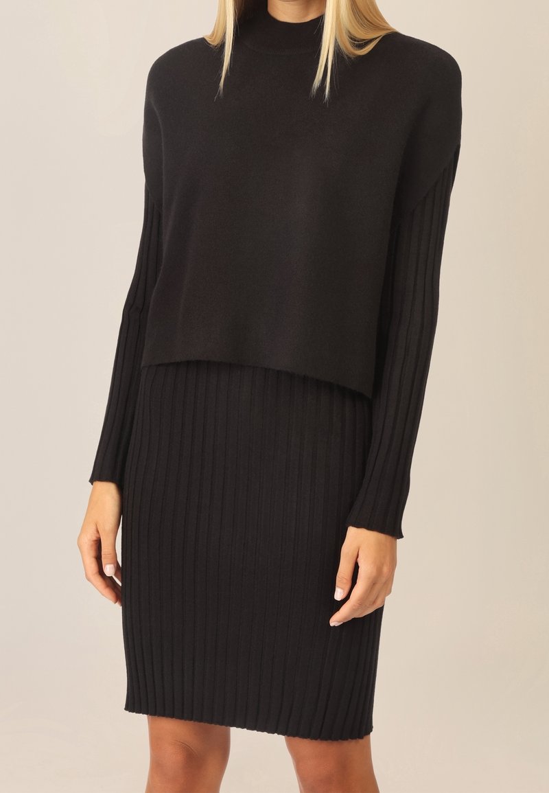Femme portant une robe en maille côtelée noire superposée à un pull noir ample à manches longues, sur un fond clair.
