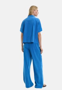 Kobieta z blond włosami, ubrana w luźną, jasnoniebieską koszulę z krótkim rękawem i szerokie spodnie, stojąca boso na tle jednolitego białego tła.