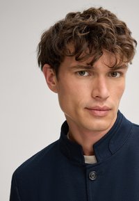 Marineblaue Jacke mit Stehkragen, strukturiertem Stoff und Knopfverschluss. Nahaufnahme eines Gesichts mit heller Haut und lockigem Haar.