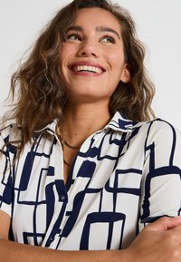 Chemise à manches courtes avec un fond blanc et des motifs rectangulaires bleu marine, un col pointu et une texture de tissu lisse.