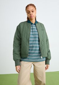 Giacca bomber verde trapuntata con polsini a coste, indossata sopra una polo a righe turchesi, abbinata a pantaloni beige a gamba larga.