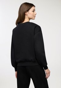 Donna con capelli castani che indossa un ampio felpa nera e pantaloni neri, rivolta di lato con la testa leggermente voltata verso la macchina fotografica.