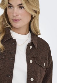 Chaqueta de mezclilla marrón con un patrón de leopardo, con cierres de botones plateados y bolsillos en el pecho, llevada sobre una camiseta blanca de cuello redondo.