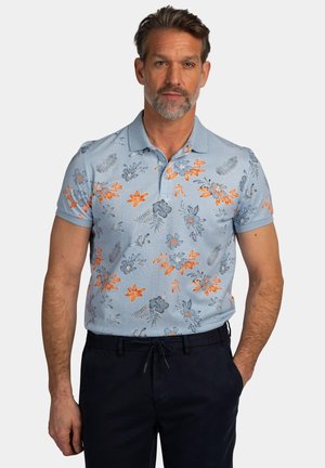 Man met grijze baard die een lichtblauw poloshirt draagt met oranje en blauwe bloemenprint en een donkere broek, staand tegen een effen achtergrond.