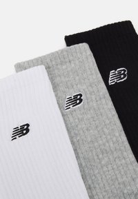 Tre par med ribbede sokker i hvit, grå og svart, hver med en liten brodert logo i hvit på oversiden.