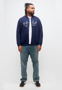 Άνδρας με dreadlocks που φοράει μπλε σκούρο μπουφάν Polo Ralph Lauren, λευκό πουκάμισο, μπλε τζιν και μαύρα αθλητικά παπούτσια, στέκεται μπροστά σε λευκό φόντο.