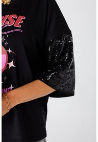 Zwarte t-shirt met een roze grafisch ontwerp, featuring zwarte korte mouwen met pailletten en een relaxte pasvorm. Textuurcontrasten tussen de materialen.