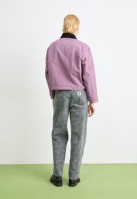 Paarse cropped jas met zwarte kraag, gecombineerd met grijze pants met een tonale bloemenpatroon en zwarte schoenen, gezien van achter.