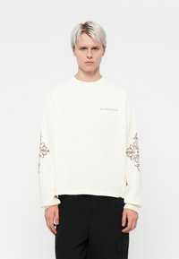Witte sweater met lange mouwen, voorzien van bruine decoratieve patronen op de mouwen, met een ronde hals en subtiele tekst met de woorden "VERSPIL CULTUUR NIET."