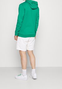 Felpa verde con tasca a marsupio e cappuccio regolabile, abbinata a pantaloni corti bianchi e sneakers bianche con dettagli verdi.