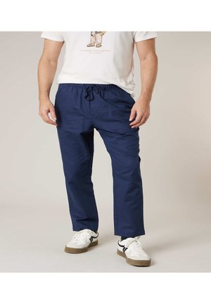 Man met witte sneakers, marineblauwe broek met trekkoord en een wit t-shirt met afbeelding, staand tegen een effen achtergrond.