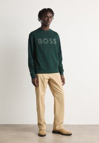 Felpa verde con logo "BOSS" grigio, abbinata a pantaloni beige e stivali in suede color tan. Il modello si trova su uno sfondo neutro.