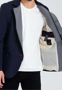 Marinblå blazer med slät yta, grårandig foder och kontrasterande bruna knapp. Inkluderar innerfickor och ytterficka med dragkedja.