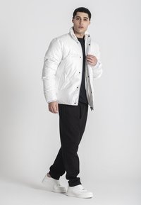 Hvid pufferjakke med en blank finish, udstyret med trykknapper, sidelommer og en opretstående krave, båret over en sort skjorte og bukser.