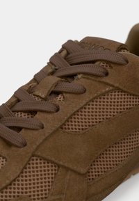 Καφέ σουέτ αθλητικό παπούτσι με πάνελ από δίχτυ, στρογγυλή μύτη και επίπεδα κορδόνια. Υφή με διακριτική λεπτομέρεια λογότυπου στην πλευρά.