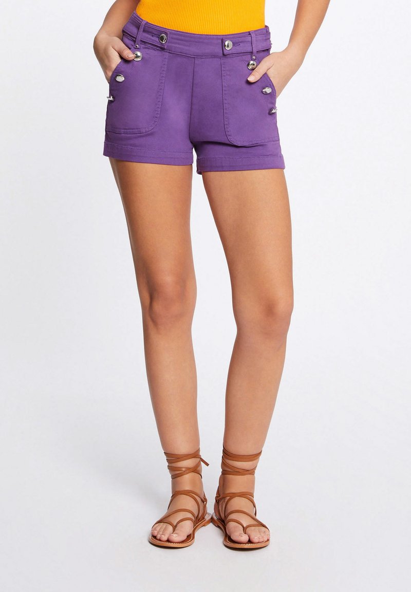 Pantaloni corti viola con due tasche frontali, dettagli con bottoni argentati e un design aderente. Abbinati a sandali marroni con cinturini.