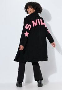 Cappotto teddy nero con testo rosa "SMILE" e dettagli a stella sulle maniche. Presenta una vestibilità rilassata e cuciture a vista. Abbinato a pantaloni neri e stivali.