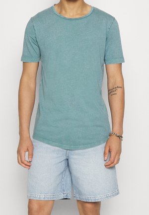 Homme portant un t-shirt turquoise à manches courtes et un short en jean bleu clair, avec un bracelet et un tatouage sur son avant-bras gauche.