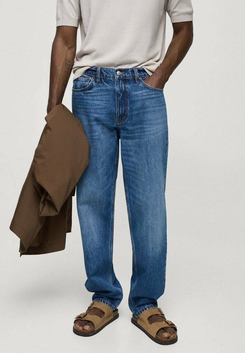 Light blue denim jeans with a relaxed fit, visible stitching, and five-pocket design. Paired with a beige shirt and brown jacket. Brown sandals.