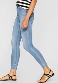 Jeans ajustados de color azul claro en tela de mezclilla con una cintura de altura media, que presentan un ligero desgastado, diseño clásico de cinco bolsillos, combinados con sandalias negras de tiras.