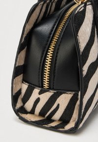 Pochette zébrée noir et beige avec un dessus en cuir noir et une fermeture éclair dorée. Texture douce, forme angulaire et détails de couture visibles.