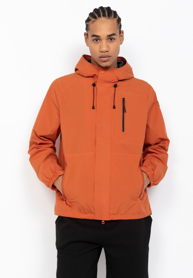 Oranje waterdichte jas met capuchon, ritszak, elastische manchet en een gladde textuur. Draag over zwarte broek op een effen achtergrond.