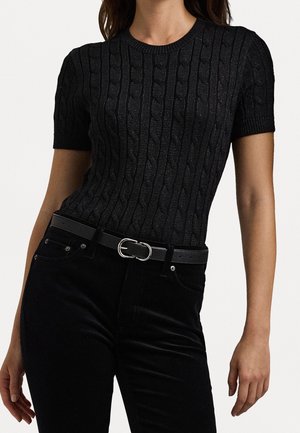 Vrouw die een aansluitende zwart kabelgebreide top met korte mouwen draagt, met zwarte broek en een zwarte riem met een zilveren dubbele ringgesp.