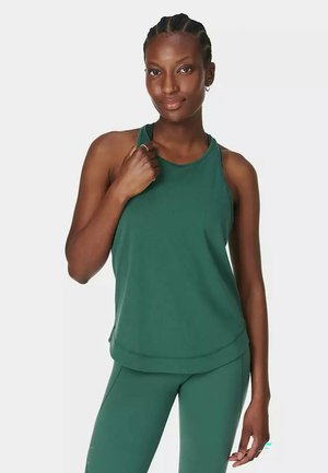 Vrouw met gevlochten haar die een groen mouwloos topje en bijpassende legging draagt, staand met één hand die de bovenste band aanraakt.