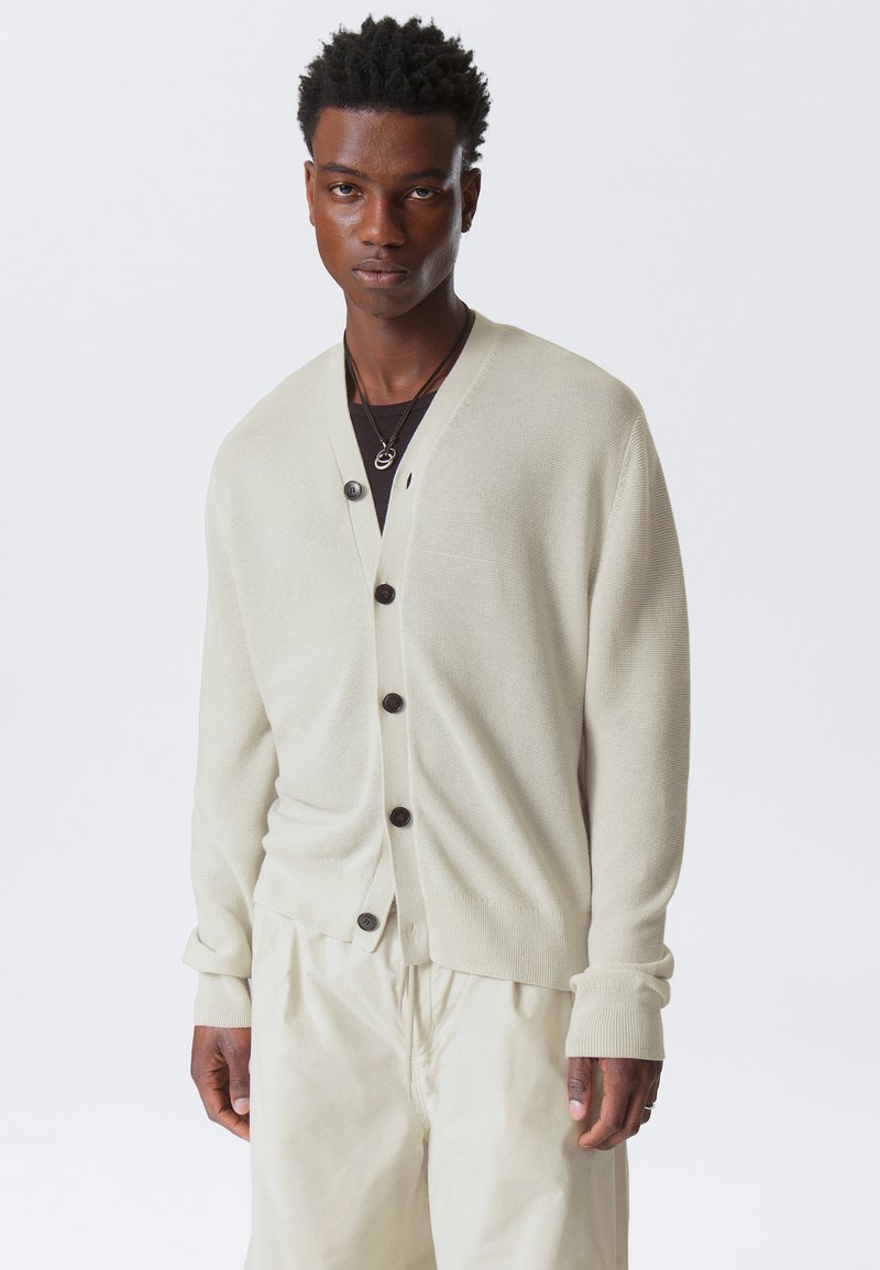 Cardigan beige clair en tissu tricoté doux, avec un col en V, cinq boutons noirs et des poignets côtelés, porté sur une chemise noire.