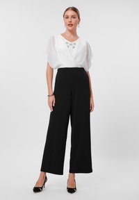 White blouse with short draped sleeves and silver embellishments, paired with high-waisted black wide-leg trousers and black heels.