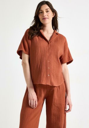 Kvinde med bølget brunt hår iført løs, struktureret, rustfarvet skjorte med korte ærmer og matchende vidde bukser mod hvid baggrund.