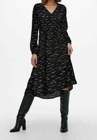 Černé šaty s dlouhými rukávy, výstřihem do V, páskem v oblasti pasu a bílými textovými vzory po celém povrchu. Stylizováno s černými kozačkami ke kolenům.