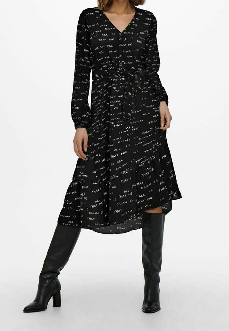 Černé šaty s dlouhými rukávy, výstřihem do V, páskem v oblasti pasu a bílými textovými vzory po celém povrchu. Stylizováno s černými kozačkami ke kolenům.