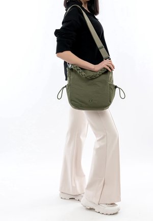 Groene schoudertas met een gevlochten handvat, twee trekkoorden en een gladde textuur. Gedragen met lichtgekleurde broek en sneakers.