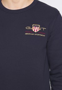 Sudadera de marina con cuello redondo, que presenta un logo bordado en oro y rojo con el texto "GANT" y "AMERICAN SPORTSWEAR". Textura suave.