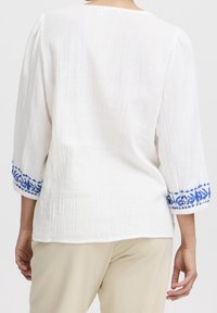 Top bianco in lino con maniche a tre quarti. La parte inferiore presenta dettagli ricamati blu con motivi floreali. Tessuto morbido e testurizzato.