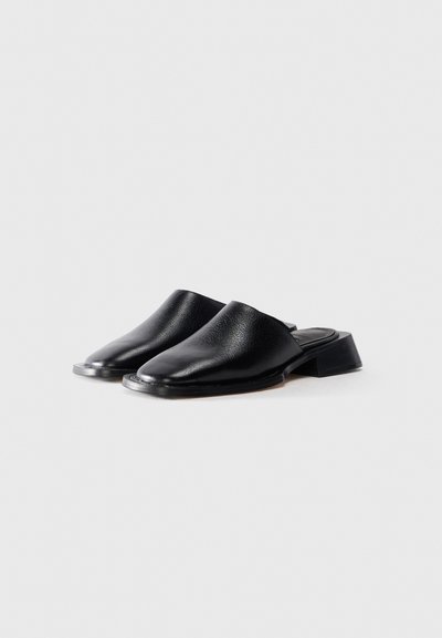 Une paire de mules en cuir noir à enfiler avec des talons blocs bas sur un fond blanc uni, orientées vers la gauche.