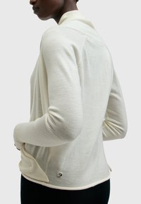 Jersey de punto beige claro con puños y dobladillo acanalados, con un brillo sutil y un pequeño adorno metálico decorativo cerca de la parte inferior.