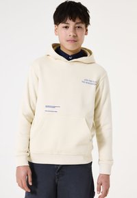 Hoodie crema realizzato in morbido tessuto, caratterizzato da una tasca a marsupio frontale, testo ricamato in blu e polsini a coste. Indossato con pantaloni corti scuri.