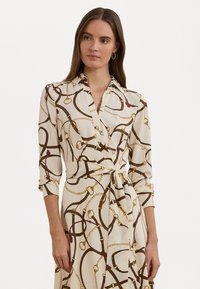 Long-sleeve wrap dress in cream fabric with brown and gold chain pattern. Features a collared neckline and tied waist for tailored fit.